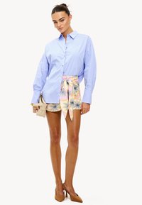 Mujer con una camisa oversized azul clara metida dentro de unos pantalones cortos florales en tonos pasteles con un cinturón atado, sujetando un clutch beige y llevando tacones beige.