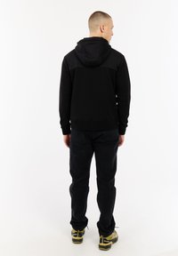 Man die met zijn rug naar de camera staat, gekleed in een zwarte capuchonjack, zwarte broek en gele en zwarte wandelschoenen, tegen een witte achtergrond.