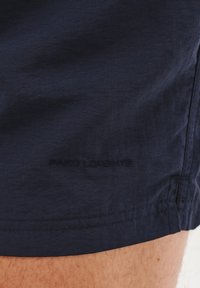 Granatowe szorty wykonane z gładkiego materiału. Posiadają subtelne logo, przeszywaną krawędź oraz elastyczny pas. Struktura powierzchni bez wzorów.