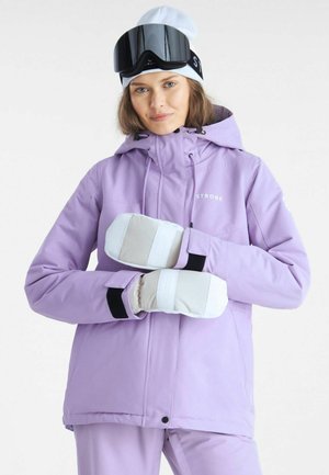 Kvinde i lavendelfarvet skijakke og bukser iført hvide luffer, hvid hue og sorte skibriller på panden, poserer med krydsede hænder.