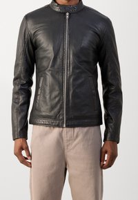 Schwarze Lederjacke mit Stehkragen, Frontreißverschluss und seitlichen Taschen. Glatte Textur und taillierte Passform, kombiniert mit hellen Hosen.