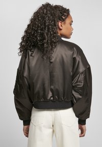 Svart satin bomberjacka med en kortad design, elastiska ärmslut och nederkant, som har en mjuk textur och en oversized passform. Vita byxor under.