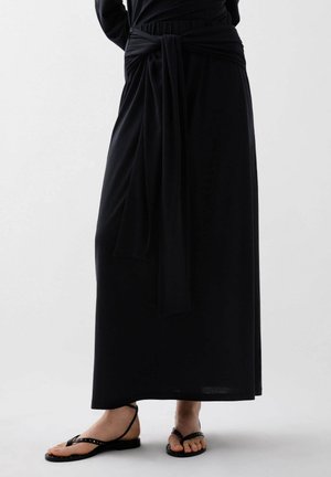 Black floor-length dress with long sleeves, featuring a tied waist detail. Paired with black flat sandals with studs along the straps.