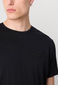 Tricou din bumbac negru, cu decolteu rotund și un logo subtil brodat pe piept, având o textură netedă și uniformă.