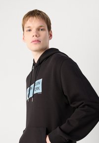 Zwarte hoodie met een voorzak, voorzien van blauwe logopatches met de tekst "Calvin Klein NEW YORK." Er zijn trekkoorden en geribbelde manchetten.