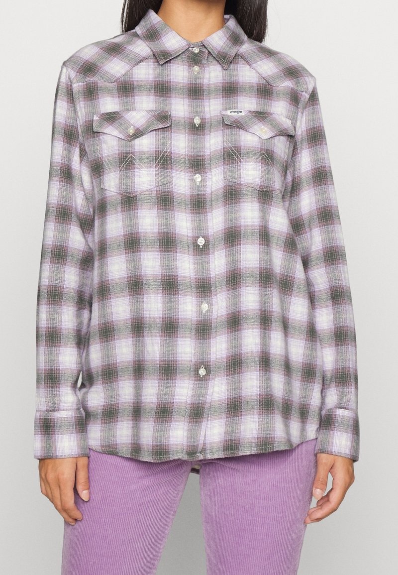 Chemise en flanelle à carreaux avec des motifs violets, verts et blancs, dotée d'un col, d'une fermeture à boutons et de deux poches poitrine. Texture de tissu douce.