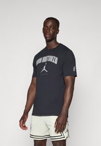 Juodas medvilnės marškinėliai su užrašu "SAN ANTONIO" ir Jumpman logotipu. Dėvimi su baltais tinkliniais šortais su juodais akcentais.