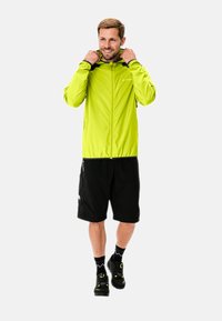Helle grüne wasserdichte Jacke mit vollem Reißverschluss und verstellbarem Kapuzen, kombiniert mit schwarzen Shorts und Schuhen mit gelben Akzenten.
