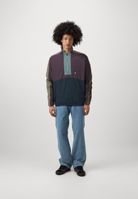 Purple and blue zip-up jacket with green accents, lightweight fabric, elastic waistband, logo patch, worn with light blue jeans and black shoes.