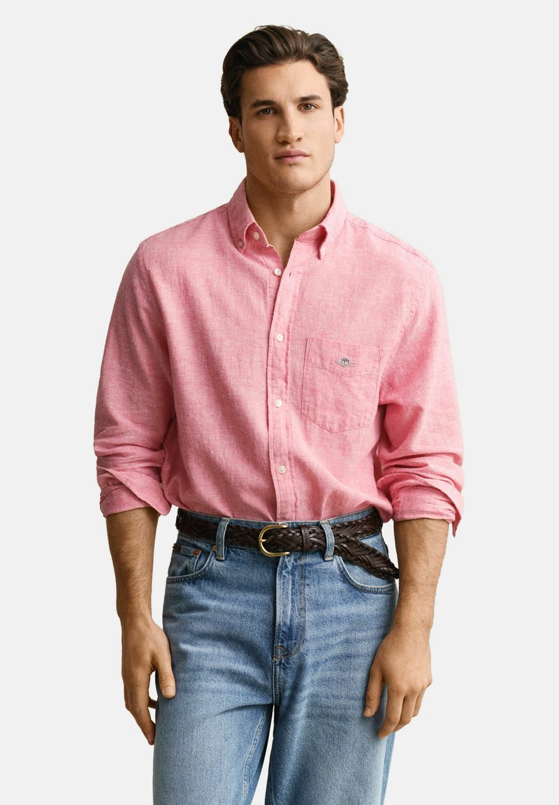 Homme portant une chemise rose à manches longues avec une poche poitrine, manches retroussées, ceinture tressée marron et jean bleu clair, sur un fond uni.