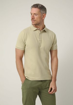 Hombre con el pelo corto que lleva una camisa polo beige y pantalones verde oliva, de pie con una mano en el bolsillo mientras mira hacia la izquierda.