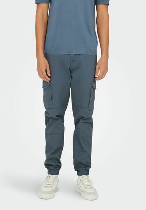 Cargo broeken van donkergrijze stof met elastische manchetten en zijzakken, gecombineerd met witte sneakers en een blauw shirt met korte mouwen.