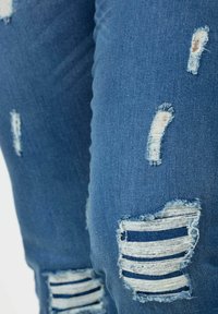 Světle modré džínové kalhoty s opotřebovanými záplatami s horizontálními pruhy a třepícími se okraji, zdůrazňujícími ležérní, potrhaný design.