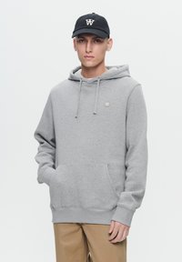 Sweat à capuche gris avec une poche frontale, fabriqué en matériau doux. Il possède des cordons de serrage et un petit logo sur la poitrine. Associé à un pantalon beige.