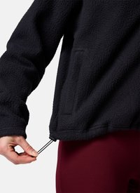 Hand die de zoom van een zwarte, getextureerde fleecejas met een elastisch koordje verstelt, draagt donkerrode broek.