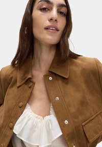Giacca in suede marrone con colletto, chiusura a bottoni e taschino sul petto, abbinata a una blusa bianca con volant sotto.