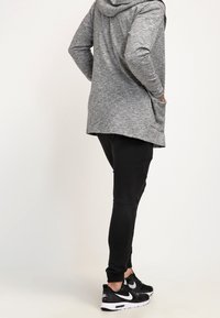 Persona che indossa un maglione grigio con cappuccio, pantaloni da jogging neri e sneakers Nike nere, in piedi con le mani nelle tasche davanti a uno sfondo semplice.