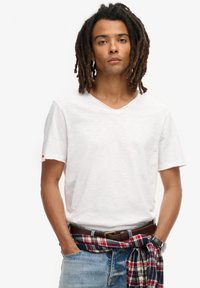 Hvid bomulds V-hals t-shirt med korte ærmer, med en slidt søm. Bæres sammen med blå denimjeans og en ternet skjorte knotet om taljen.