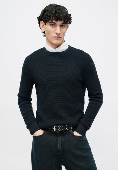 Marineblauwe ribgebreide trui met ronde hals. Gecombineerd met donkerblauwe spijkerbroek en een zwarte riem met zilveren oogjes. Model staat met handen in de zakken.