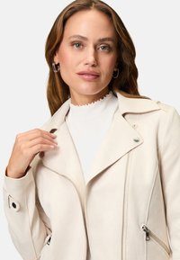 Giacca in ecopelle color crema con zip frontale, colletto angolato e dettagli in metallo color argento. Presenta tasche laterali con zip e una texture liscia.