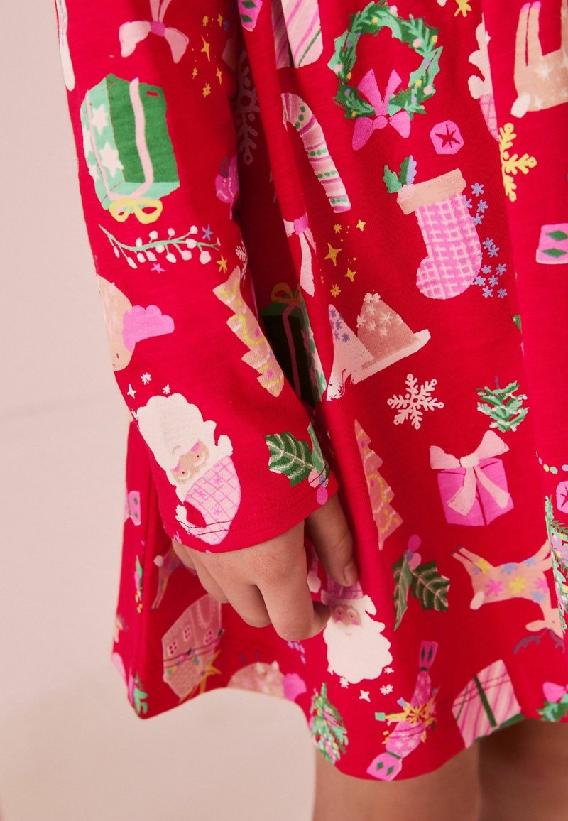 Main d'enfant et robe rouge avec des motifs de Noël colorés, y compris le Père Noël, des cadeaux, des couronnes, des bas, et des cannes de bonbon.