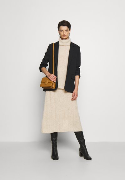 Blazer preto sobre um vestido de gola alta creme, combinado com botas pretas até ao joelho e uma pequena mala cruzada castanha. Texturas suaves e forma ajustada.