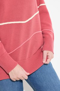 Koronkowy sweter w kolorze koralowym z kontrastowymi białymi poziomymi paskami, z żeberkowymi mankietami i dołem, założony na niebieskie dżinsy. Miękka faktura i luźny krój.