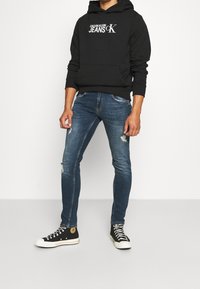 Felpa nera con cappuccio e logo bianco "Calvin Klein Jeans"; jeans blu a taglio slim e strappati; sneakers nere alte con suola bianca.