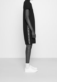 Svart polo klänning med korta ärmar som har sidomärken i vit text. Bärs med vita sneakers och ankelsockar för en avslappnad look.