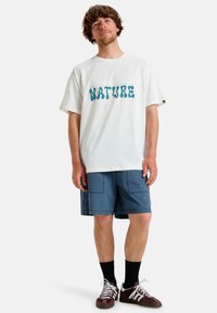 Balta medvilninė marškinė su mėlynu "NATURE" grafiniu dizainu. Suderinta su mėlynomis šortais, juodomis kojinėmis ir rudais sportbačiais su baltais dryžiais.