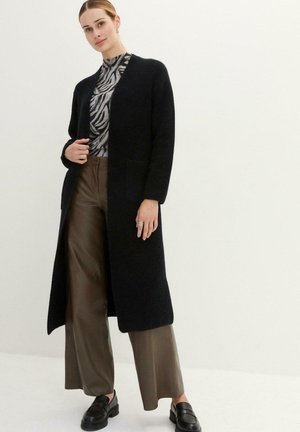 Long cardigan noir à l'avant ouvert, associé à un haut à imprimé animal gris et à un pantalon plissé marron, porté avec des mocassins noirs.