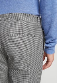 Kalhoty s černobílým vzorem houndstooth a hladkým pasem, se dvěma zadními kapsami, spárované se světle modrým svetrem.