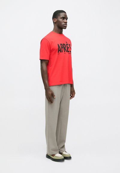 Άνδρας στέκεται, φορώντας κόκκινη μπλούζα με το κείμενο "APRÈS", μπεζ παντελόνι με φαρδιά πόδια και δύο τόνους πράσινα και καφέ παπούτσια, ενάντια σε λευκό φόντο.