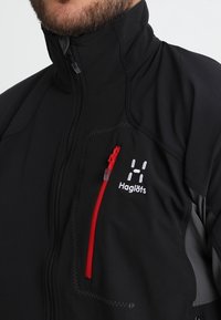 Svart mjukskaljacka med en röd dragkedjeficka, vitt logotyp och grå detaljer. Hög krage med dragkedja längs hela framsidan.