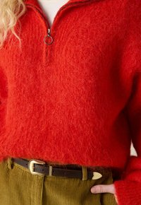 Roter, flauschiger Pullover mit halbem Reißverschluss und Metallring, kombiniert mit olivgrünen Cordhosen und einem schwarzen Gürtel mit goldener Schnalle.