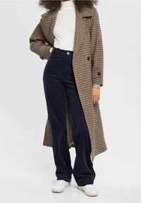 Długi brązowy płaszcz w kratę noszony na białym golfie, zestawiony z granatowymi spodniami z kordurowej tkaniny i białymi trampkami. Posiada duże guziki i boczne rozcięcia.