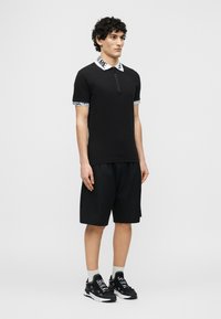 Άνδρας που στέκεται, φορώντας μαύρο κοντομάνικο πουκάμισο με φερμουάρ και λευκό κείμενο στον γιακά, μαύρο σορτς μέχρι το γόνατο, λευκές κάλτσες και μαύρα αθλητικά παπούτσια με λευκό κείμενο.