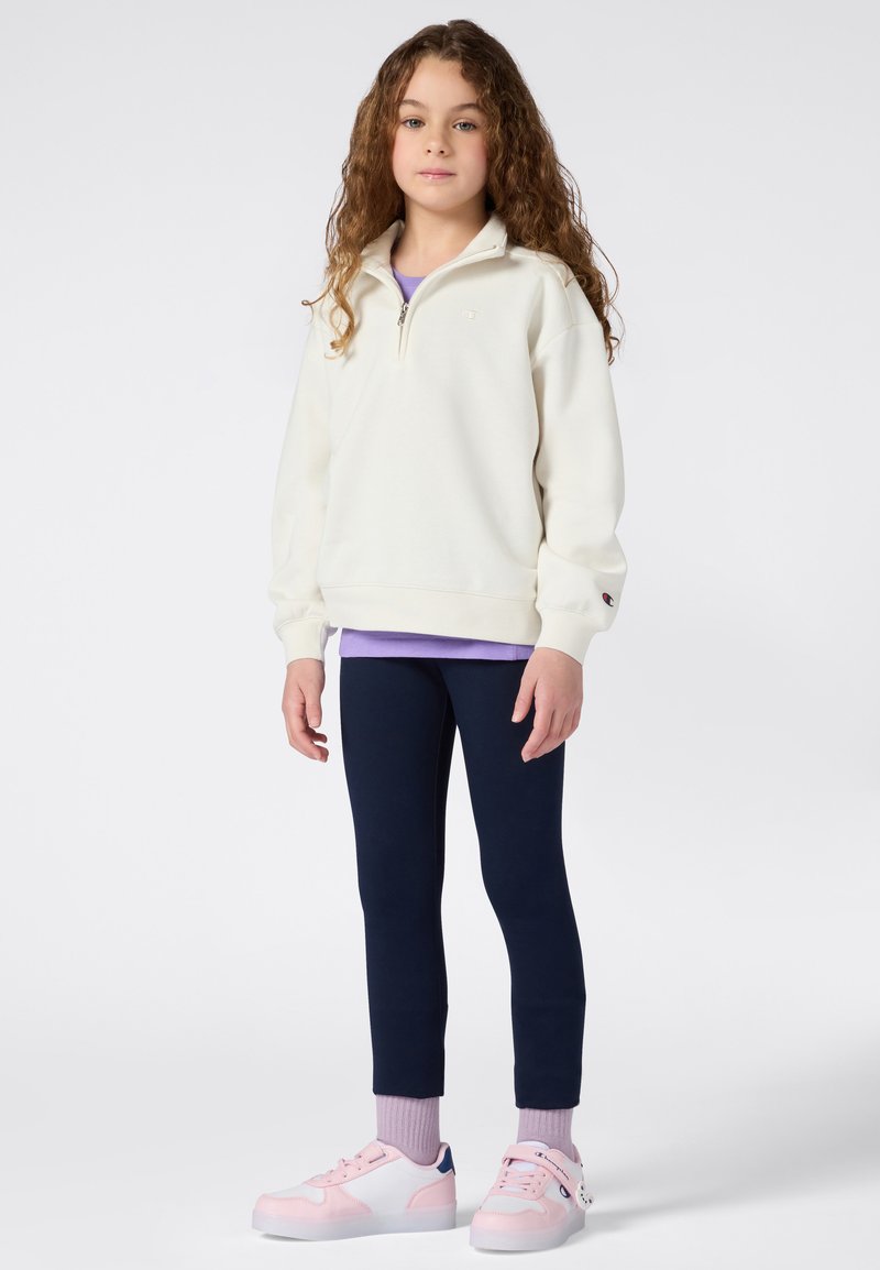 Sudadera blanca con cremallera, camiseta de color púrpura debajo, leggings de color azul marino, zapatillas rosas y blancas con un acento azul, de pie contra un fondo liso.