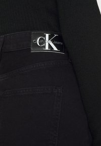 El bolsillo trasero de unos vaqueros negros con una visible etiqueta de cuero de Calvin Klein Jeans y la manga de un suéter negro de canalé al lado.