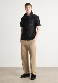 Czarna koszula z krótkim rękawem z kieszenią, beżowe spodnie i czarne buty. Na koszuli jest zarzucony sweter na ramionach. Prosty design.