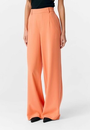 Frau trägt hoch taillierte, weit geschnittene Pfirsichfarbene Hose kombiniert mit schwarzen spitzen Schuhen und einem schwarzen ärmellosen Oberteil vor weißem Hintergrund.