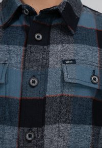 Koszula flanelowa w kratę w odcieniach niebieskiego, szarości i czerni z pomarańczowymi akcentami. Posiada zapięcie na guziki oraz kieszenie na piersi. Teksturowany materiał.