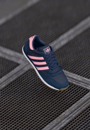 Granatowo-różowy sneaker Adidas z trzema paskami, sznurowadła zawiązane, spoczywający na przemysłowej metalowej kratce podłogowej.