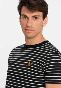 Černobíle pruhované tričko s žebrovaným dekoltem a malou zlatou emblémem na hrudi. Krátké rukávy s přiléhavým střihem.