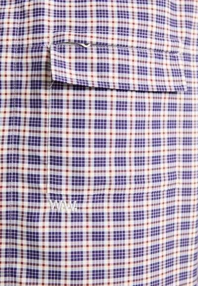 Tejido de cuadros en tonos azul, rojo y crema. Presenta un bolsillo de parche y detalles bordados. Textura ligera y suave.