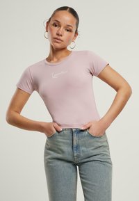 Lys rosa tettsittende t-skjorte med korte ermer, rund hals og hvit skriftlogo på brystet. Båret med høyt liv blå denim jeans.