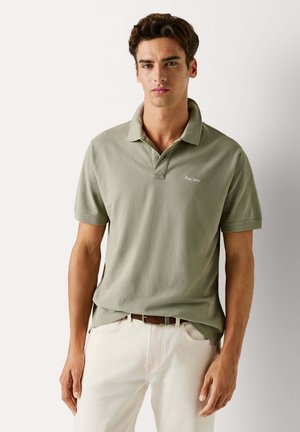 Zaļgani olīvu polo krekls ar apkakli, īsām piedurknēm un izšūtu logotipu kreisajā krūtīs; saskaņots ar gaiši krāsām džinsiem.