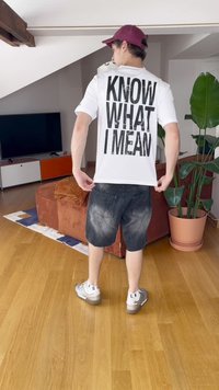 Bela majica s velikim črnim besedilom "VEŠ, KAJ MISLIM" na hrbtni strani, v kombinaciji s temno denim kratkimi hlačami in sivo obutvijo na leseni tla.