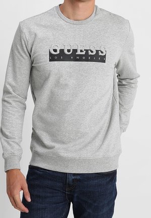 Mann trägt ein hellgraues Sweatshirt mit langen Ärmeln und dem Logo "GUESS Los Angeles" sowie eine dunkelblaue Jeans, steht vor einem einfarbigen Hintergrund.