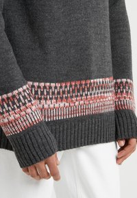 Pull en maille grise avec un poignet côtelé et un motif bicolore en corail et blanc au bas, mettant en valeur un détail soigné.